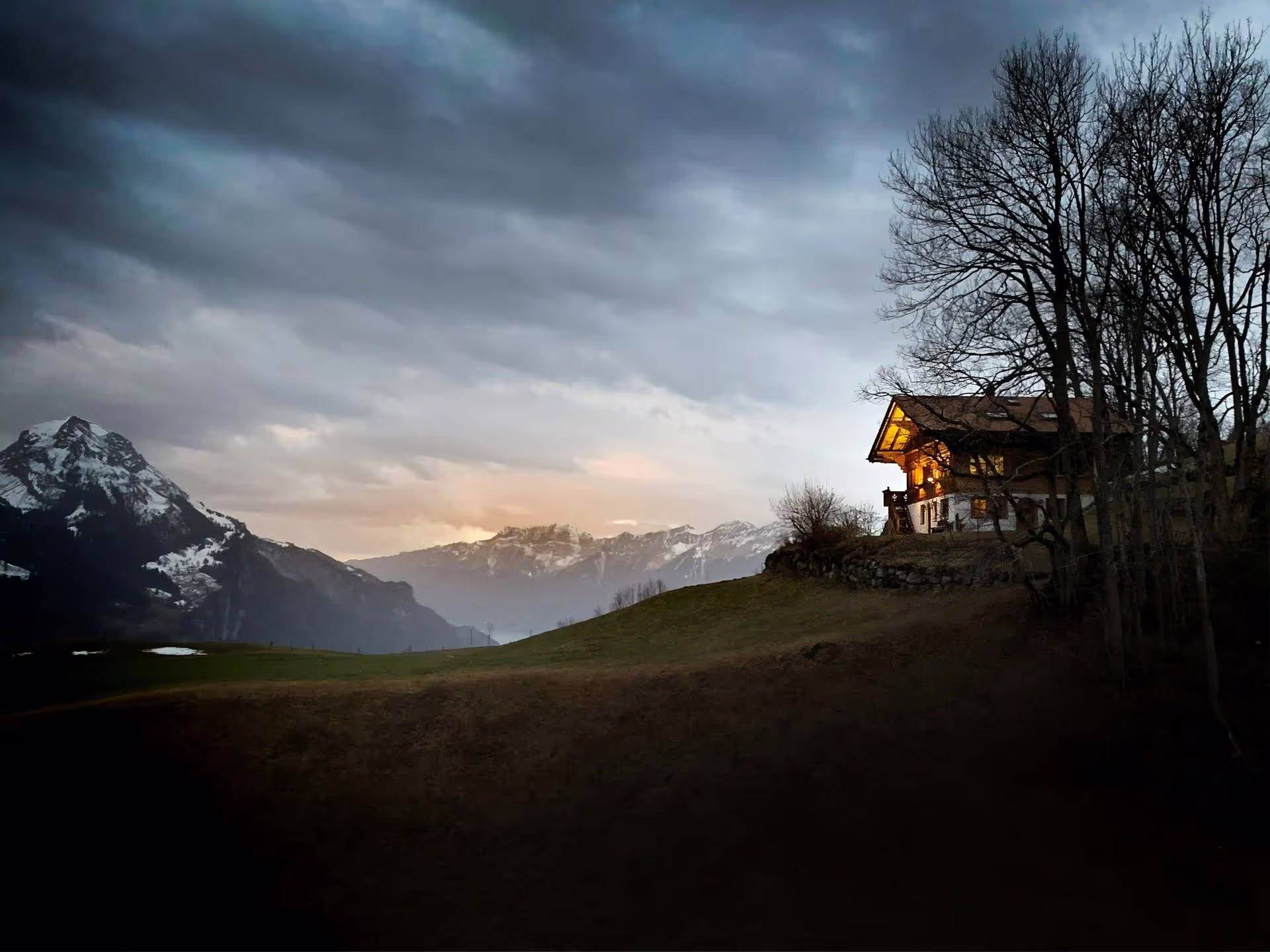 A cozy mountain house illuminated at dusk, surrounded by snow-capped peaks and dark, moody skies.